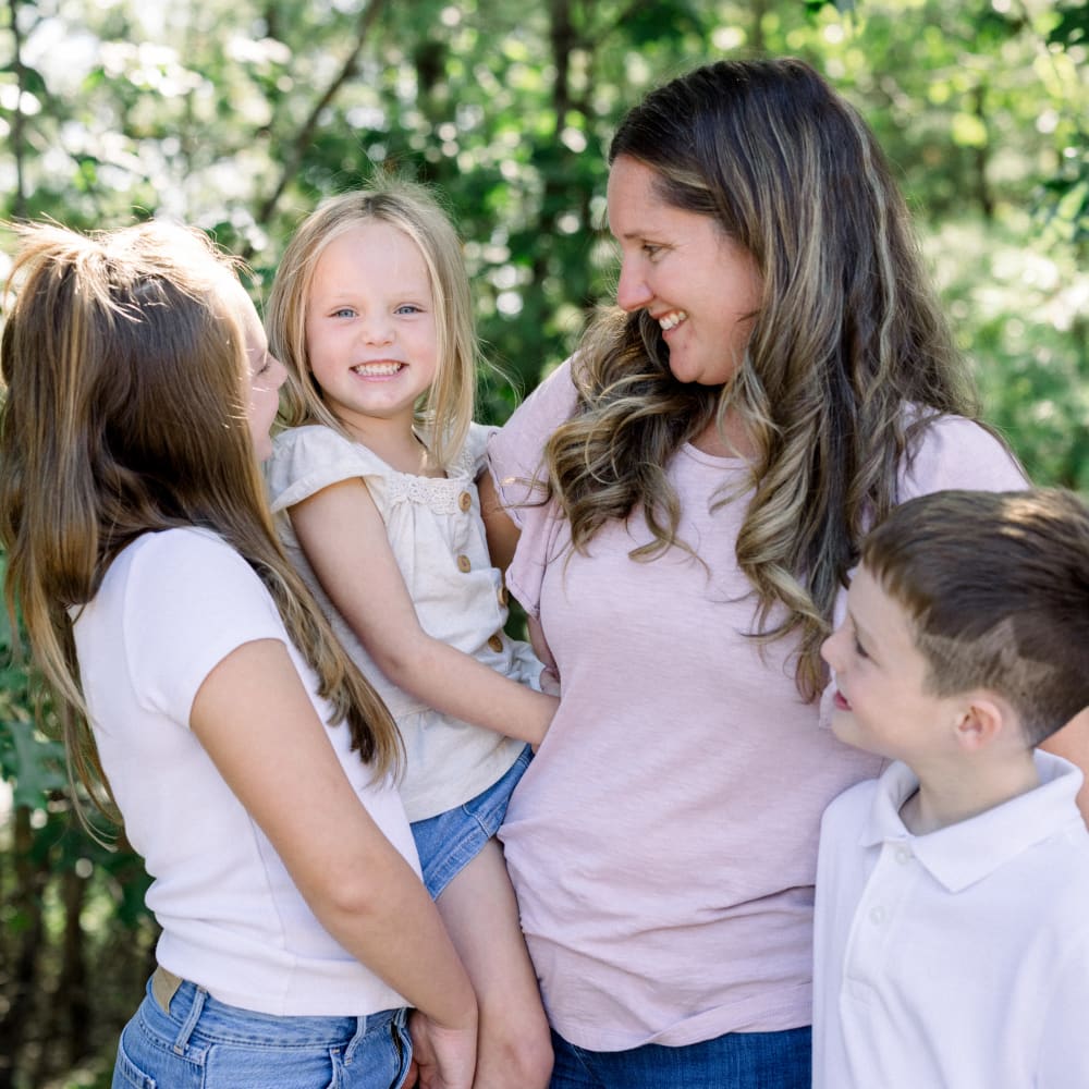 A woman and her three children are all smiling at each other. She is holding her youngest child, a blond girl, who is looking directly at the camera.