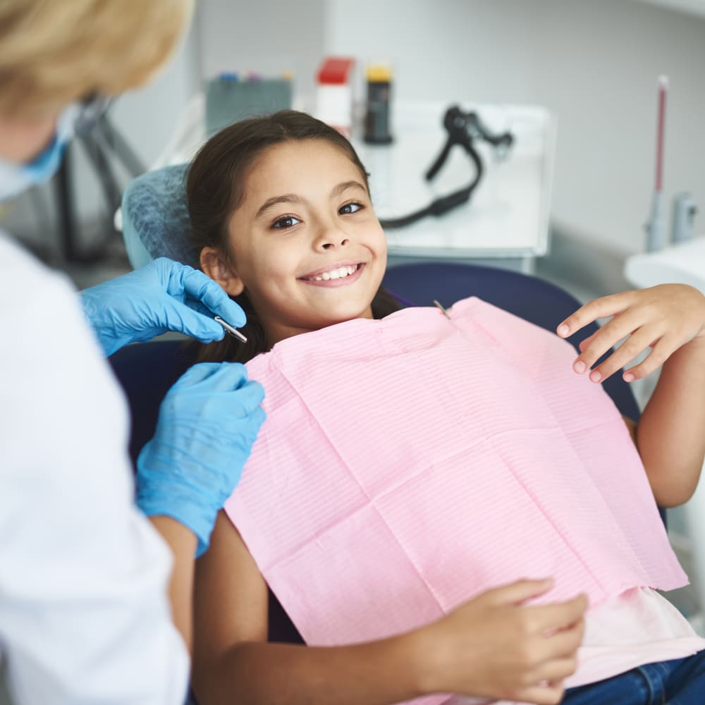 A young girl sits in a dentists chair. She is wearing a pink cloth on her chest, and is smiling at the camera.