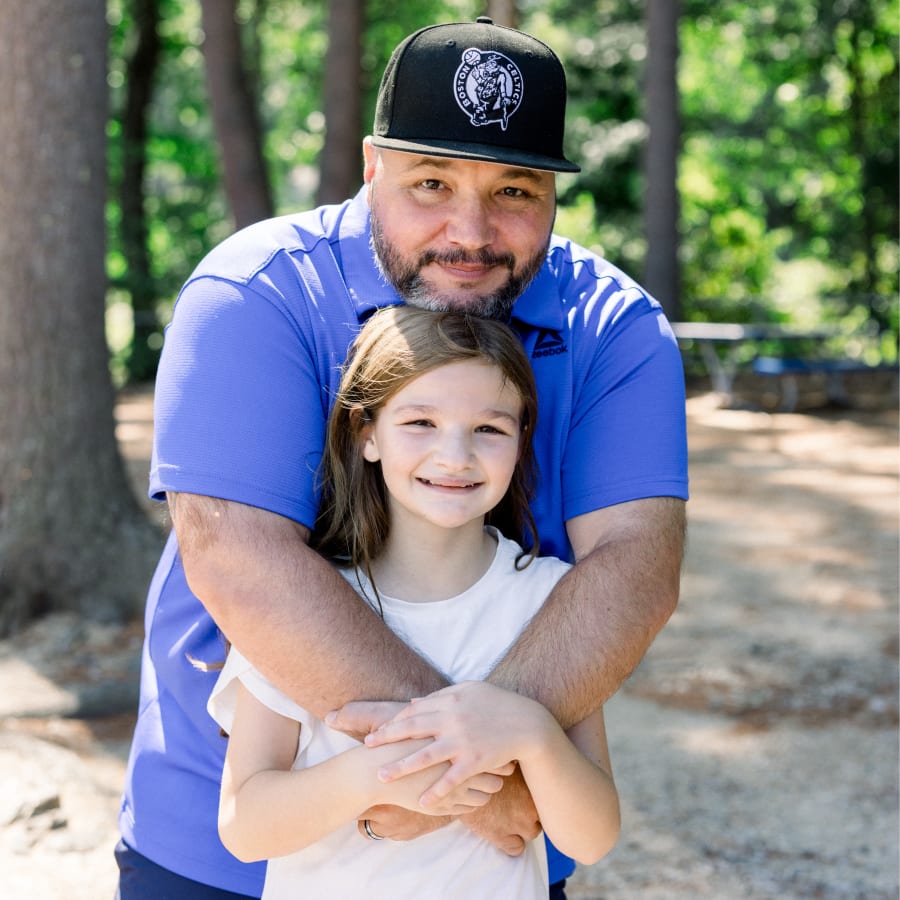 A man is standing behind a young girl with his arms wrapped around her and his head resting on her head. She is smiling and holding his hands.