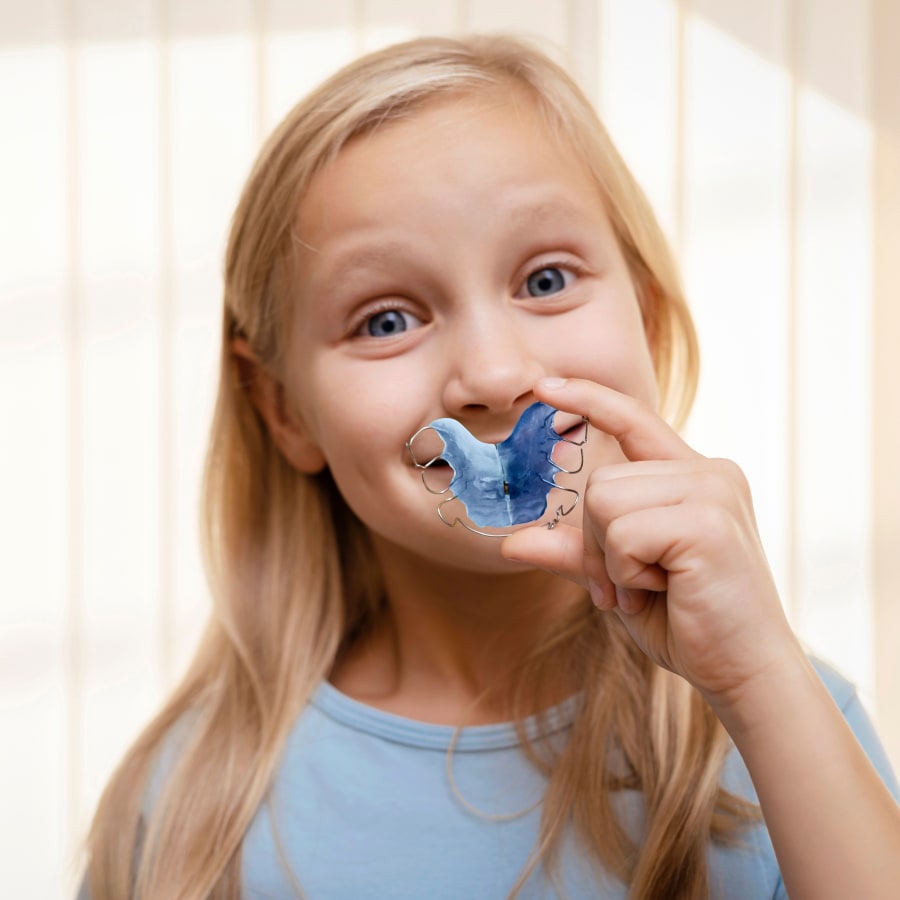 A young blonde girl is holding up a dental retainer in front of her mouth.