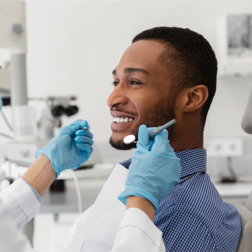 An adult man is sitting in a dental chair smiling. Only the dentist's gloved hands are in frame, as they approach him with tools. 