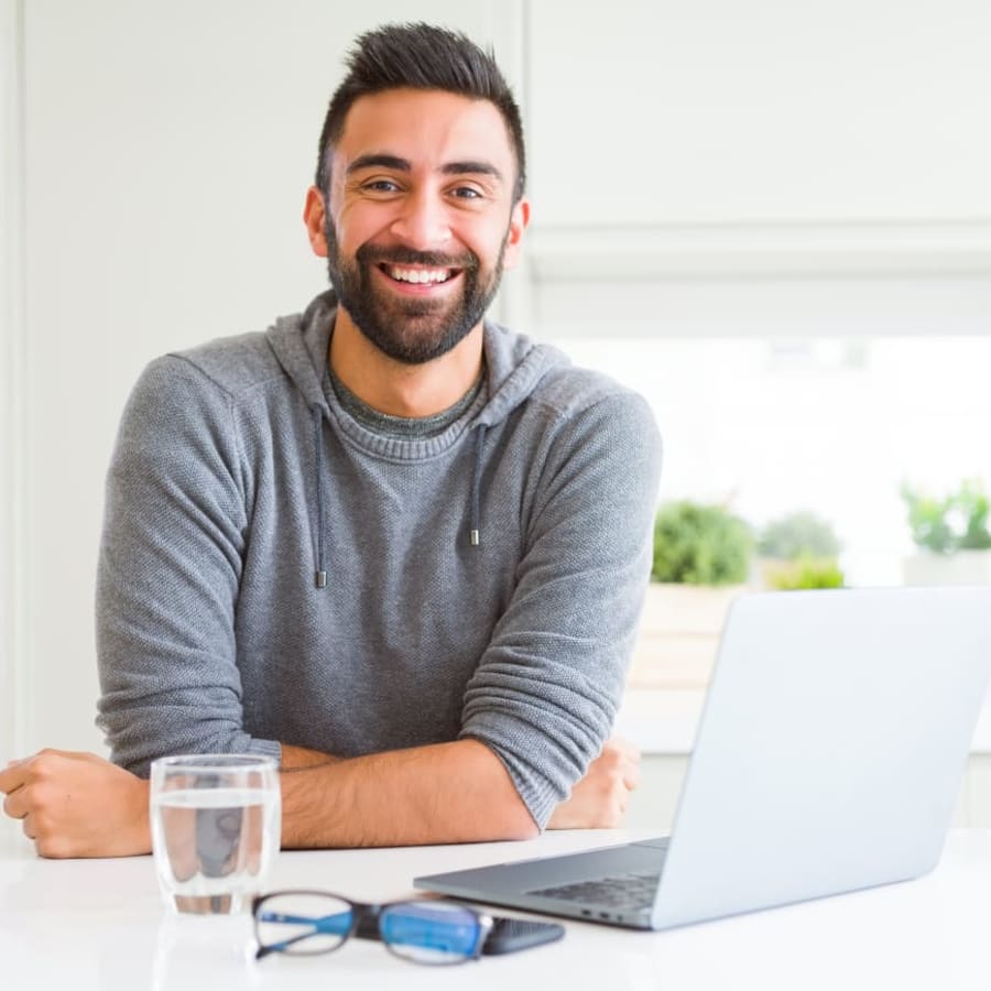 A man is smiling and leaning on a desk. There is a laptop in front of him and a class of water.