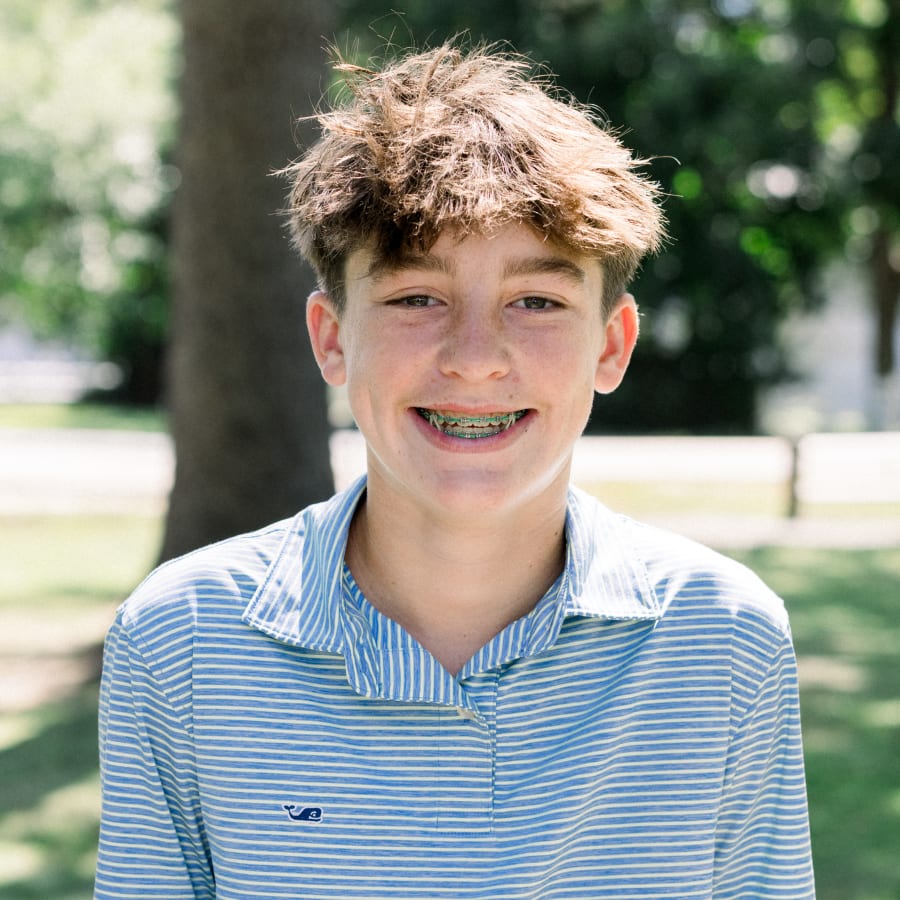 A teen boy in a blue and white horizontal striped shirt is sitting outside smiling, showing off his green braces. On his braces, he also has elastics.
