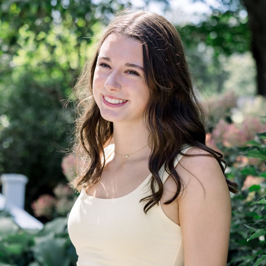 A teenage girl is wearing a yellow dress and smiling. She is standing outside near some foliage.